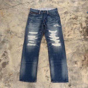 May include: Blue denim jeans with distressed detailing. The jeans feature multiple ripped sections with white backing, a zipper pocket, and a classic five-pocket design. The jeans are laid flat on a concrete surface.