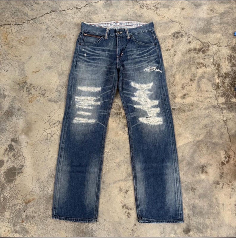 May include: Blue denim jeans with distressed detailing. The jeans feature multiple ripped sections with white backing, a zipper pocket, and a classic five-pocket design. The jeans are laid flat on a concrete surface.