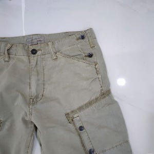 May include: Close-up of khaki-colored cargo shorts. The shorts feature multiple pockets, button closures, and a textured fabric. The design includes reinforced stitching and a classic, utilitarian style.