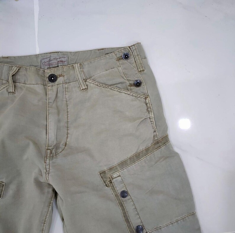 May include: Close-up of khaki-colored cargo shorts. The shorts feature multiple pockets, button closures, and a textured fabric. The design includes reinforced stitching and a classic, utilitarian style.