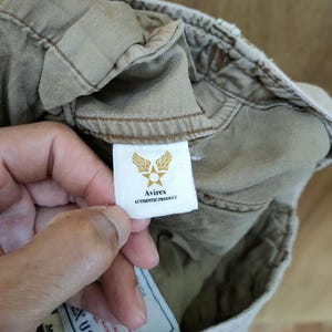 May include: Close-up of a beige garment with a white tag featuring the Avirex logo: a star within wings. The tag reads "Avirex AUTHENTIC PRODUCT". The garment's fabric is visible, showing stitching and texture.
