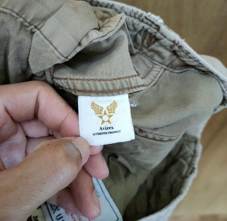 May include: Close-up of a beige garment with a white tag featuring the Avirex logo: a star within wings. The tag reads "Avirex AUTHENTIC PRODUCT". The garment's fabric is visible, showing stitching and texture.