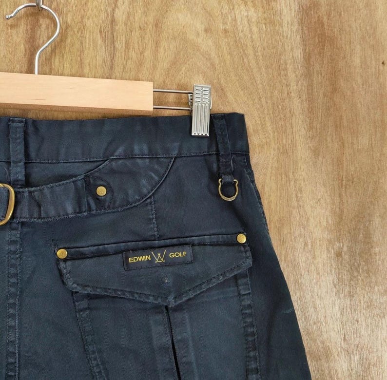 Pu&ograve; includere: Primo piano di pantaloni blu navy con una tasca con la scritta "EDWIN GOLF". I pantaloni hanno dettagli dorati e sono appesi a un'appendiabiti di legno.