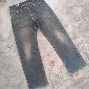 May include: Faded blue denim jeans with a distressed look. The jeans have a classic five-pocket design, a button and zipper closure, and a small tear on the right knee. The jeans are slightly worn and have a vintage aesthetic.