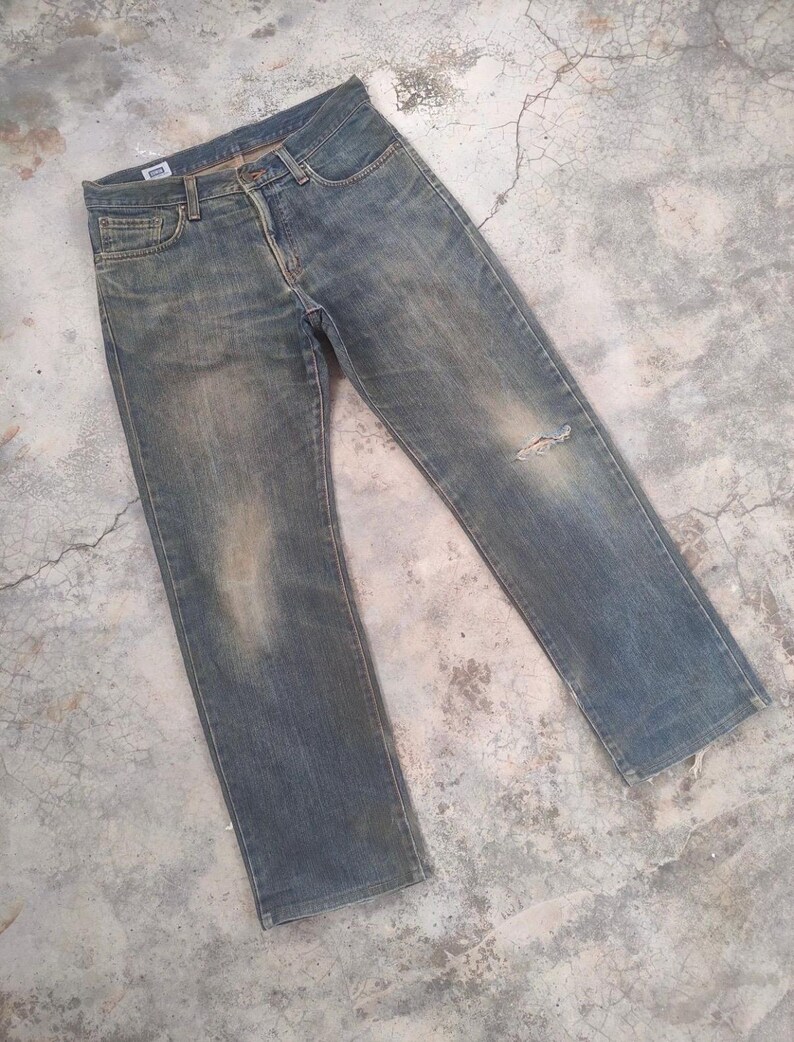 May include: Faded blue denim jeans with a distressed look. The jeans have a classic five-pocket design, a button and zipper closure, and a small tear on the right knee. The jeans are slightly worn and have a vintage aesthetic.