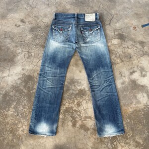 May include: Faded blue denim jeans with decorative back pockets and a small tag. The jeans have a distressed look with faded areas and visible wear patterns. The back pockets have a unique design with red accents.
