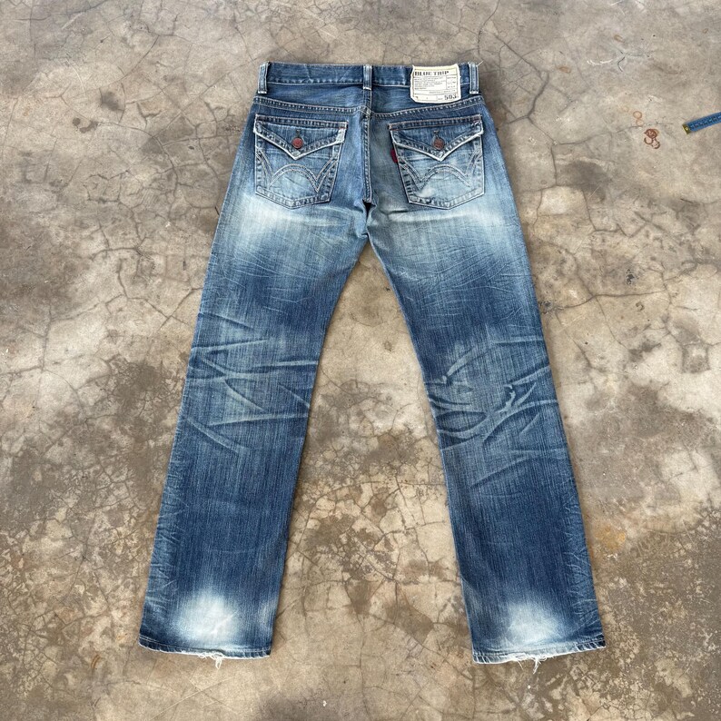 May include: Faded blue denim jeans with decorative back pockets and a small tag. The jeans have a distressed look with faded areas and visible wear patterns. The back pockets have a unique design with red accents.