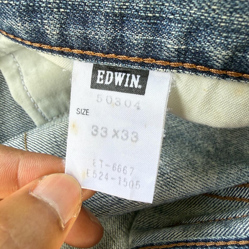 May include: Close-up of a denim jeans tag with the brand name "EDWIN" in white on a black rectangle. The tag also shows the size "33 x 33". The jeans are blue.