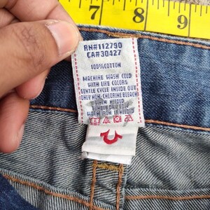 May include: A close-up of a denim jeans label with text. The white label has black text, including "RN#112790 CA#30427" and washing instructions. The jeans are blue.