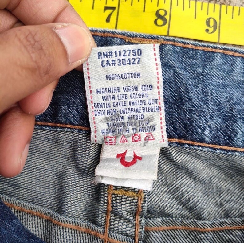 May include: A close-up of a denim jeans label with text. The white label has black text, including "RN#112790 CA#30427" and washing instructions. The jeans are blue.