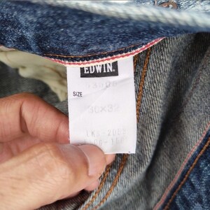 May include: Close-up of a pair of blue denim jeans with a white tag. The tag reads "EDWIN" in black letters. The size is 30x32 inches. The jeans have visible stitching and a red and white trim.