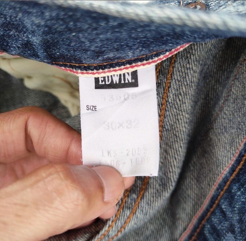 May include: Close-up of a pair of blue denim jeans with a white tag. The tag reads "EDWIN" in black letters. The size is 30x32 inches. The jeans have visible stitching and a red and white trim.