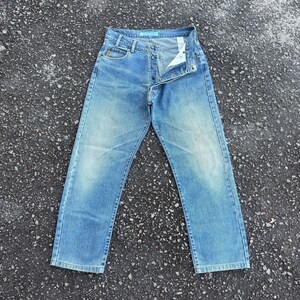 May include: Light blue denim jeans with a button fly and classic five-pocket design. The jeans have a faded wash and are laid flat on a textured surface. The waist band has a light blue tag.