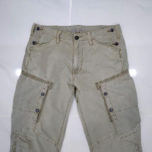 May include: Khaki-colored cargo pants with multiple pockets and button details. The pants have a faded appearance and a relaxed fit. The design includes reinforced stitching and a classic button and zipper closure. The pants are made of a durable fabric.