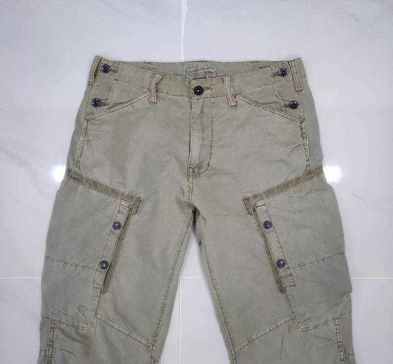 May include: Khaki-colored cargo pants with multiple pockets and button details. The pants have a faded appearance and a relaxed fit. The design includes reinforced stitching and a classic button and zipper closure. The pants are made of a durable fabric.