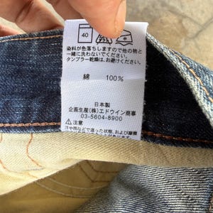 May include: Close-up of a white clothing tag with Japanese text and symbols, attached to blue denim fabric. The tag indicates the fabric is 100% cotton and provides washing instructions. The tag also includes a phone number.