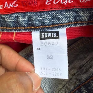 May include: Close-up of a white tag with the brand name "EDWIN" in black. The tag also displays "EG503", "SIZE", and the number "32". The tag is attached to blue denim jeans with a red interior.