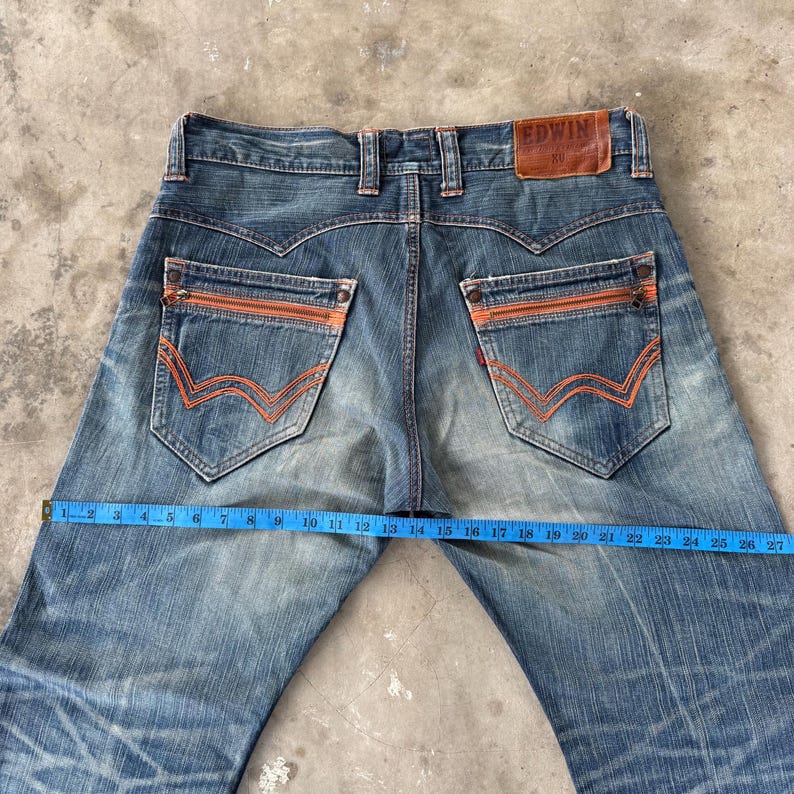 Pu&ograve; includere: Jeans in denim blu con cuciture e dettagli con cerniera arancioni sulle tasche posteriori. I jeans hanno un lavaggio sbiadito e una toppa in pelle con il marchio "EDWIN". La vita misura circa 38 cm.