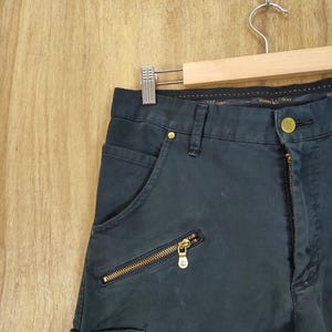 Pu&ograve; includere: Pantaloni blu navy scuro con cerniere e bottoni color oro. I pantaloni hanno una tasca con cerniera e chiusura con bottone. I pantaloni sono appesi a un'appendiabiti di legno.