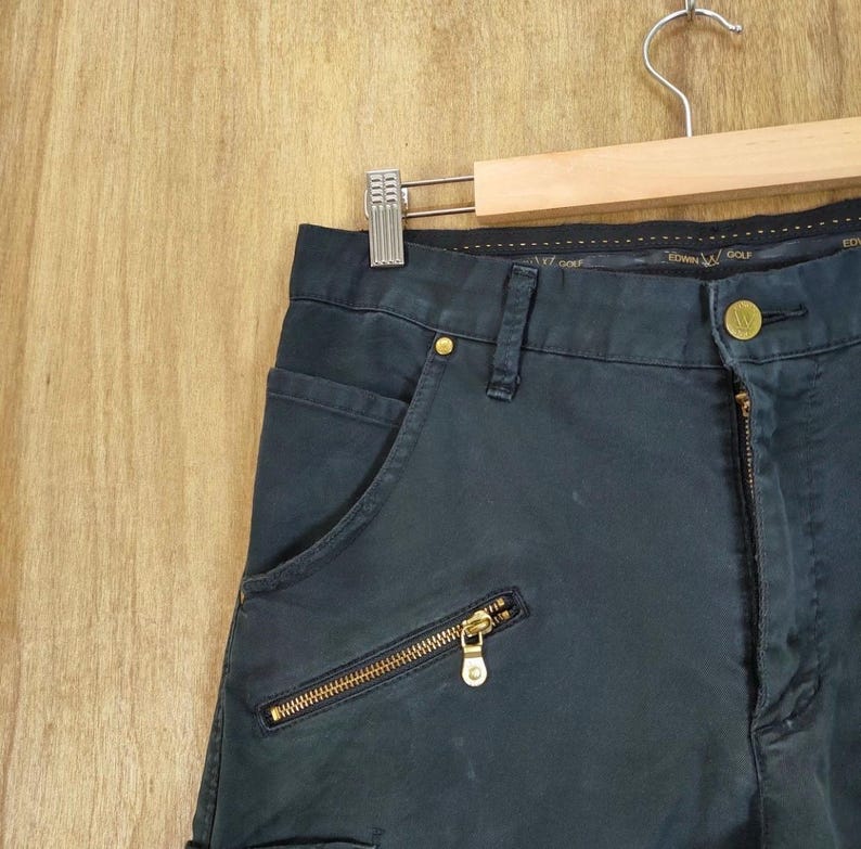 Pu&ograve; includere: Pantaloni blu navy scuro con cerniere e bottoni color oro. I pantaloni hanno una tasca con cerniera e chiusura con bottone. I pantaloni sono appesi a un'appendiabiti di legno.