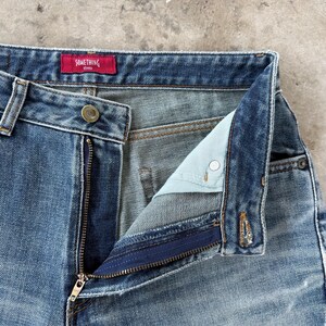 May include: Close-up of blue denim jeans with an open zipper. The jeans have a button closure and a red tag that reads "SOMETHING" in white letters. The jeans are faded and have a classic five-pocket design.