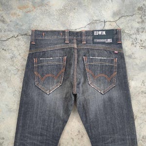 Pu&ograve; includere: Jeans in denim grigio scuro con cuciture arancioni e dettagli usurati. Le tasche posteriori presentano un design curvo distintivo. Il marchio "EDWIN" &egrave; visibile su una toppa nera in vita. I jeans hanno un classico design a cinque tasche.