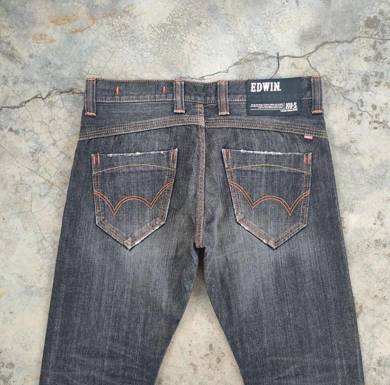 Pu&ograve; includere: Jeans in denim grigio scuro con cuciture arancioni e dettagli usurati. Le tasche posteriori presentano un design curvo distintivo. Il marchio "EDWIN" &egrave; visibile su una toppa nera in vita. I jeans hanno un classico design a cinque tasche.