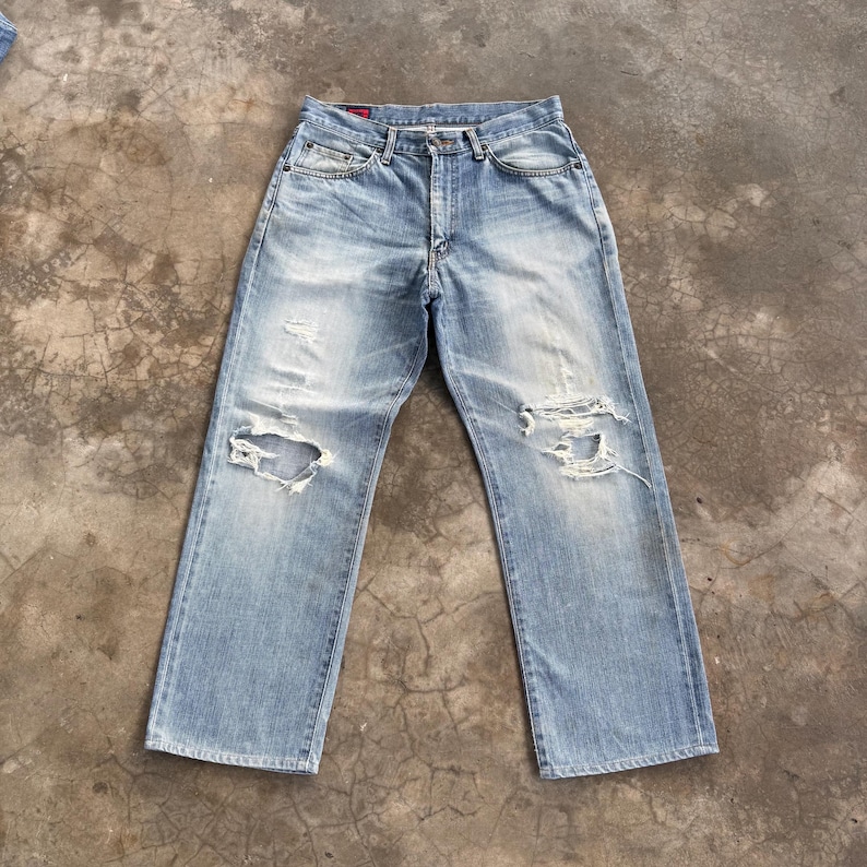 May include: Light blue denim jeans with distressed details, including rips on both knees. The jeans have a classic five-pocket design and a button and zipper closure. The denim has a faded wash.