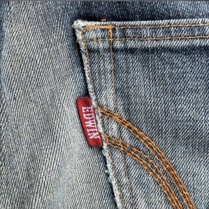 Pu&ograve; includere: Primo piano di una sezione di jeans blu, che mostra una tasca posteriore con cuciture marroni. Un'etichetta rossa con il marchio "EDWIN" &egrave; cucita sulla tasca.