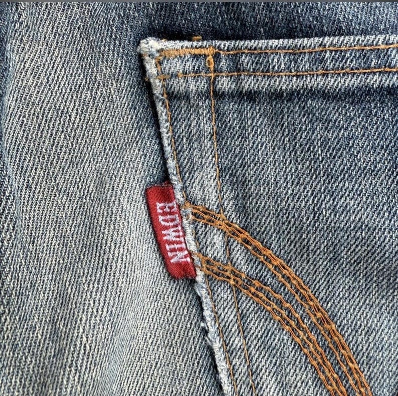 Pu&ograve; includere: Primo piano di una sezione di jeans blu, che mostra una tasca posteriore con cuciture marroni. Un'etichetta rossa con il marchio "EDWIN" &egrave; cucita sulla tasca.