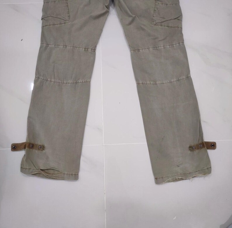 May include: Khaki-colored cargo pants with multiple pockets and stitched details. The pants have reinforced knee panels and adjustable straps at the ankles. The fabric appears to be a durable, medium-weight material.