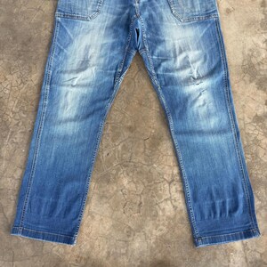 May include: Faded blue denim jeans with a classic five-pocket design. The jeans have a relaxed fit and show signs of wear, including distressed areas on the knees. The denim is a medium wash with lighter fading in areas.