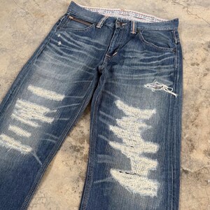 May include: Faded blue denim jeans with distressed detailing. The jeans feature multiple areas of intentional wear and tear, with large patches of exposed white fabric. The jeans have a classic five-pocket design and a zip fly.