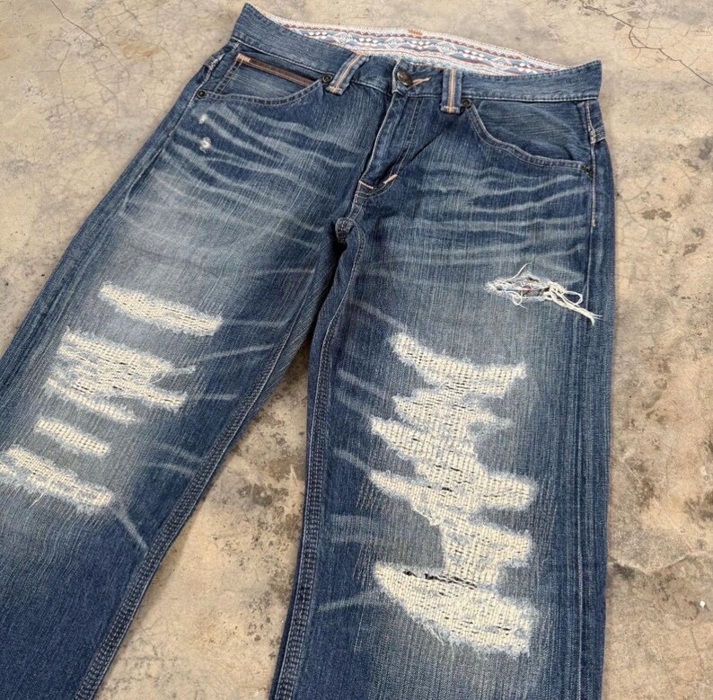 May include: Faded blue denim jeans with distressed detailing. The jeans feature multiple areas of intentional wear and tear, with large patches of exposed white fabric. The jeans have a classic five-pocket design and a zip fly.