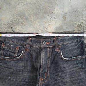 Pu&ograve; includere: Jeans in denim blu scuro con cuciture e dettagli arancioni. Un metro a nastro &egrave; appoggiato sulla vita dei jeans. I jeans hanno tasche anteriori e chiusura con bottoni. I jeans sono di uno stile classico.