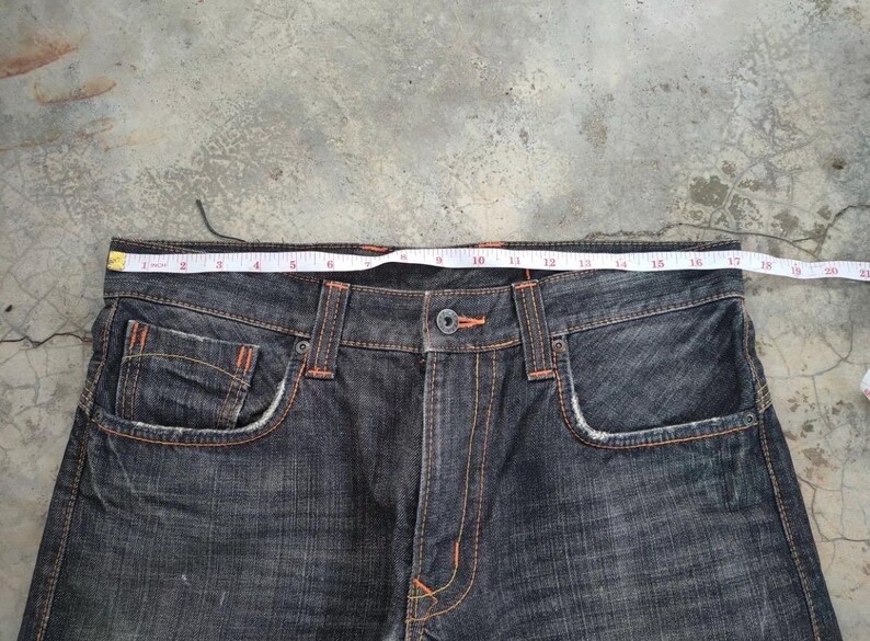 Pu&ograve; includere: Jeans in denim blu scuro con cuciture e dettagli arancioni. Un metro a nastro &egrave; appoggiato sulla vita dei jeans. I jeans hanno tasche anteriori e chiusura con bottoni. I jeans sono di uno stile classico.