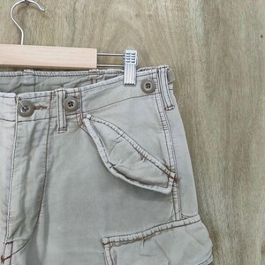 May include: Close-up of khaki cargo shorts with visible stitching and button details. The shorts are hanging on a wooden hanger with a metal clip. The fabric appears to be a durable cotton blend.