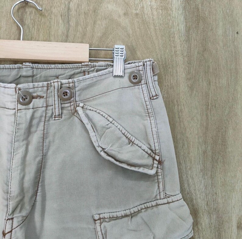 May include: Close-up of khaki cargo shorts with visible stitching and button details. The shorts are hanging on a wooden hanger with a metal clip. The fabric appears to be a durable cotton blend.