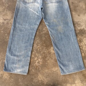 May include: Light blue denim jeans with a faded wash. The jeans have a classic five-pocket design and a straight leg cut. The denim shows signs of wear and tear, adding to its vintage aesthetic.