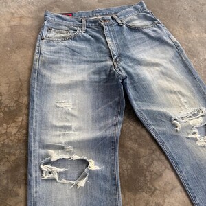 May include: Light blue denim jeans with distressed details, including rips and faded areas. The jeans have a classic five-pocket design and a button and zipper closure. The denim has a worn appearance.