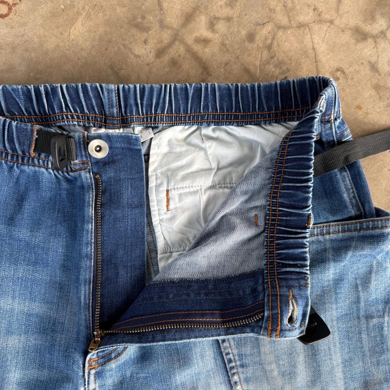 May include: Close-up of blue denim pants with an elastic waistband and a zipper. The pants have a button closure and a black belt loop. The interior lining is white. The denim has a faded appearance.