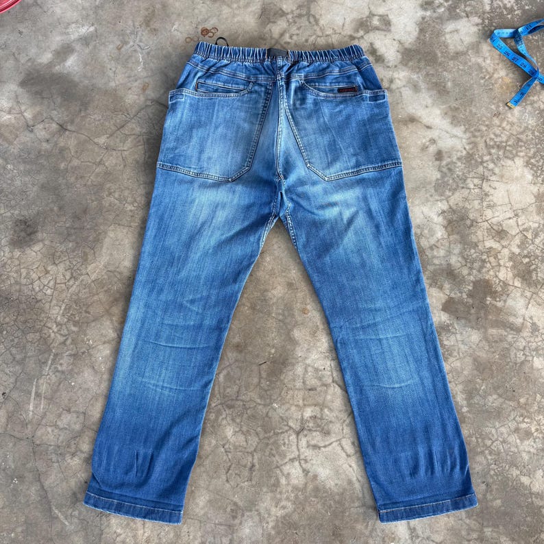May include: Back view of blue denim jeans with an elastic waistband. The jeans have two back pockets and a unique design on the back. The denim has a faded appearance.