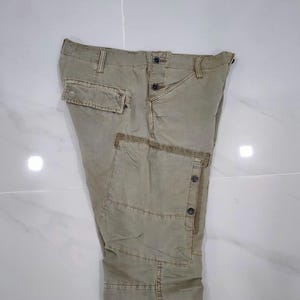May include: Khaki-colored cargo pants with multiple pockets and button closures. The pants have a relaxed fit and a slightly faded appearance, suggesting a worn-in style. The design includes reinforced stitching and a classic cargo aesthetic.
