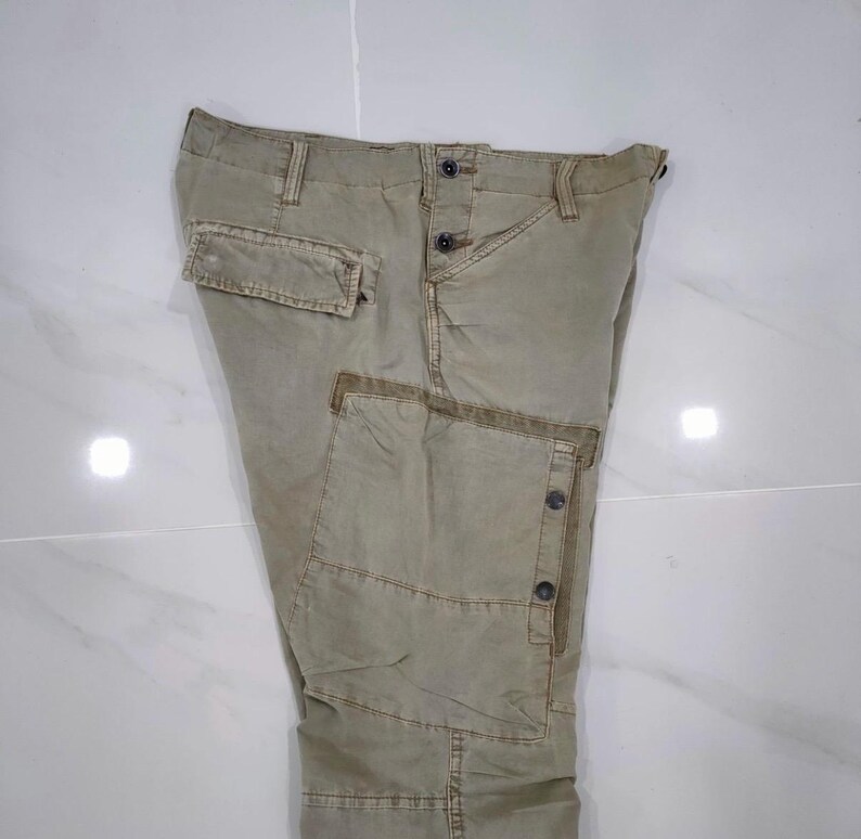 May include: Khaki-colored cargo pants with multiple pockets and button closures. The pants have a relaxed fit and a slightly faded appearance, suggesting a worn-in style. The design includes reinforced stitching and a classic cargo aesthetic.