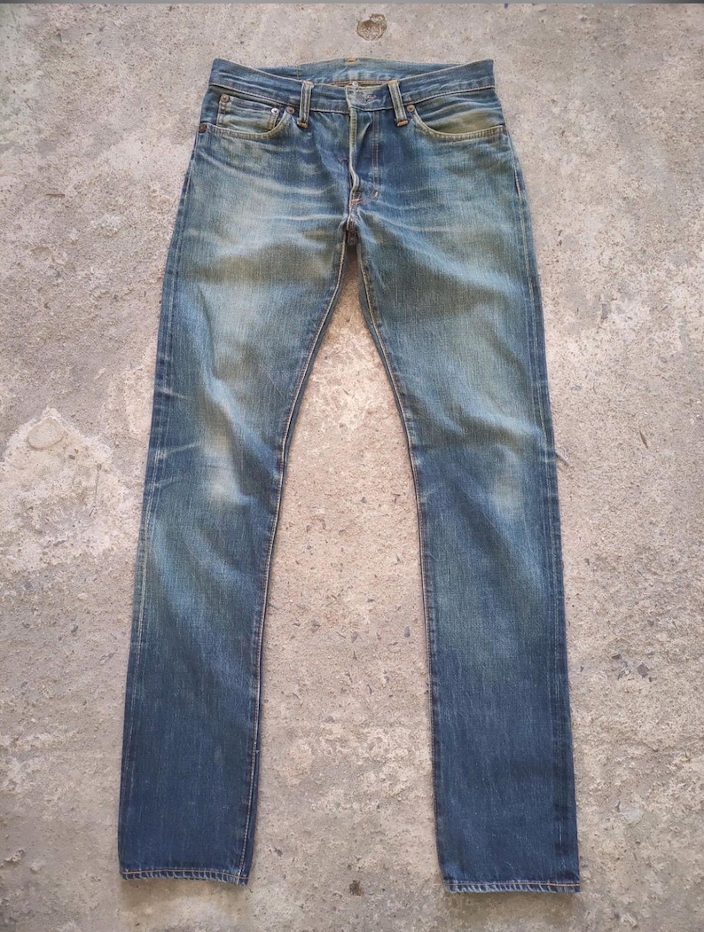 May include: Faded blue denim jeans with a button fly and classic five-pocket design. The jeans show signs of wear and fading, with a straight leg cut. The denim has a vintage, worn-in look.