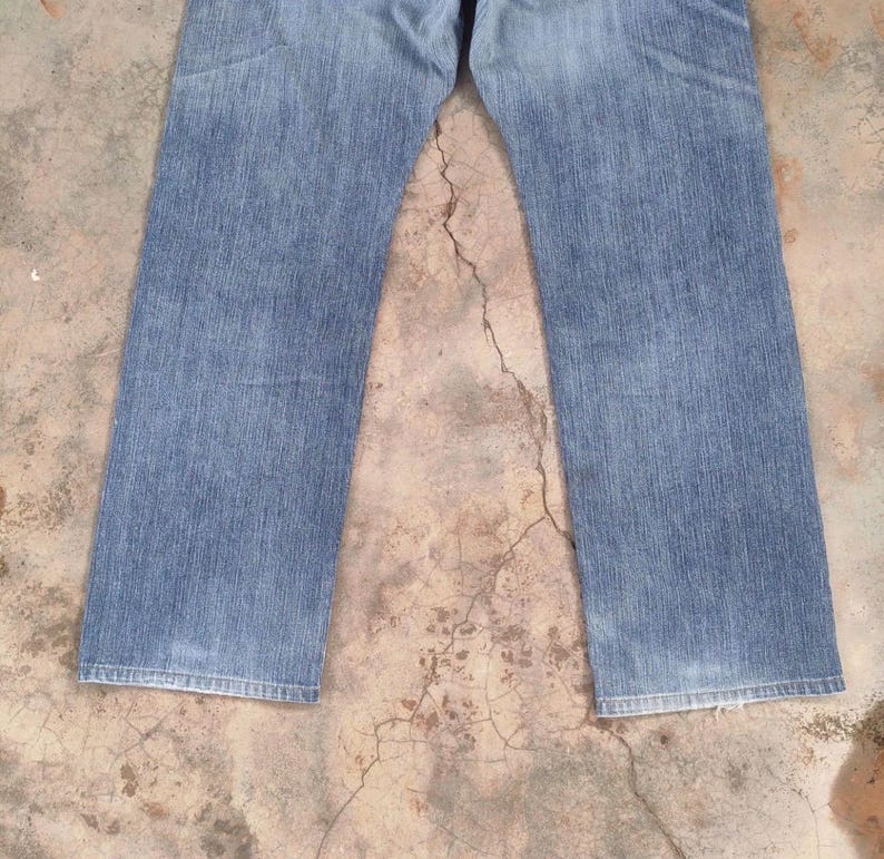 May include: Light blue denim jeans with a classic straight leg cut. The jeans show signs of wear and fading, with a medium wash. The image is a top-down view of the jeans.