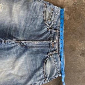 May include: Light blue denim jeans with a distressed look, featuring a classic five-pocket design. A blue measuring tape shows a length of approximately 17 inches. The jeans have a button closure and belt loops.