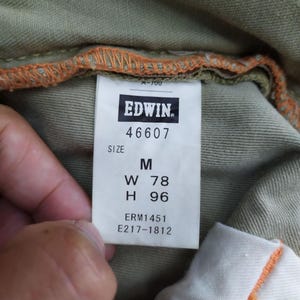 May include: Close-up of a tag from Edwin jeans. The tag is white with black text, displaying the brand name, size M, and measurements: W 78 inches, H 96 inches. The tag is sewn into olive green fabric.