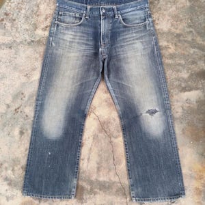 Puede incluir: Vaqueros de mezclilla azul descolorido con un aspecto desgastado. Los vaqueros tienen un dise&ntilde;o cl&aacute;sico de cinco bolsillos, bragueta con botones y un peque&ntilde;o desgarro en la rodilla derecha. La mezclilla tiene un aspecto desgastado con zonas m&aacute;s claras en los muslos y rodillas.