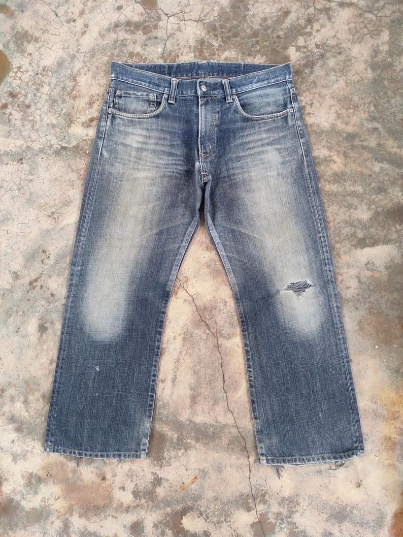 Puede incluir: Vaqueros de mezclilla azul descolorido con un aspecto desgastado. Los vaqueros tienen un dise&ntilde;o cl&aacute;sico de cinco bolsillos, bragueta con botones y un peque&ntilde;o desgarro en la rodilla derecha. La mezclilla tiene un aspecto desgastado con zonas m&aacute;s claras en los muslos y rodillas.